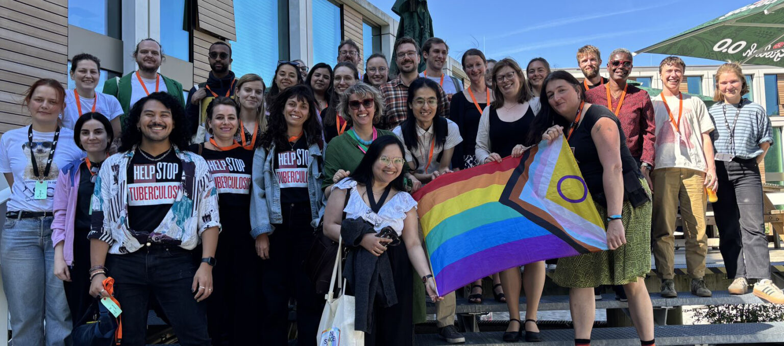 A group picture taken at the previous iteration of this event in Nijmegen. Participants are standing on some stairs, and in the front an Intersex-Inclusive Progress Pride Flag is being held.